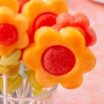 Several fruit kabobs with cantaloupe and watermelon flowers arranged in a clear glass cup.