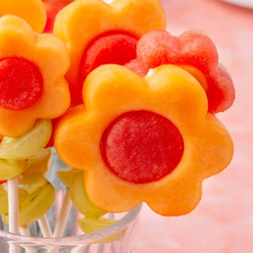 Several fruit kabobs with cantaloupe and watermelon flowers arranged in a clear glass cup.