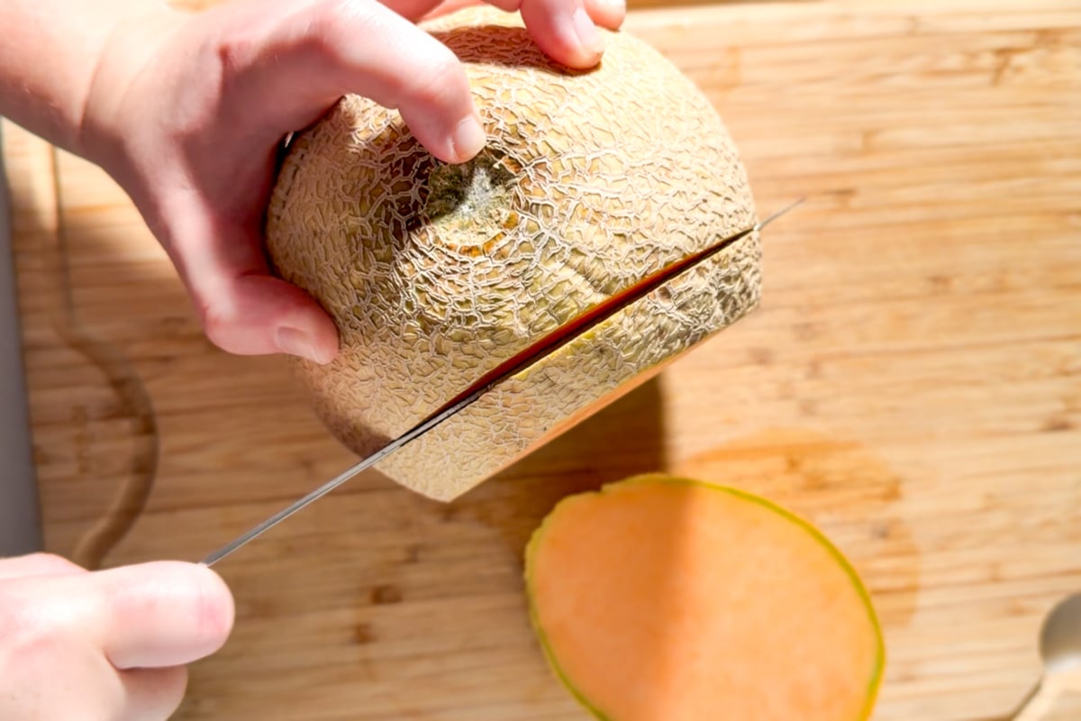 Hands slicing a cantaloupe crosswise from the side on a wooden cutting board to create seed-free rounds for fruit kabobs.