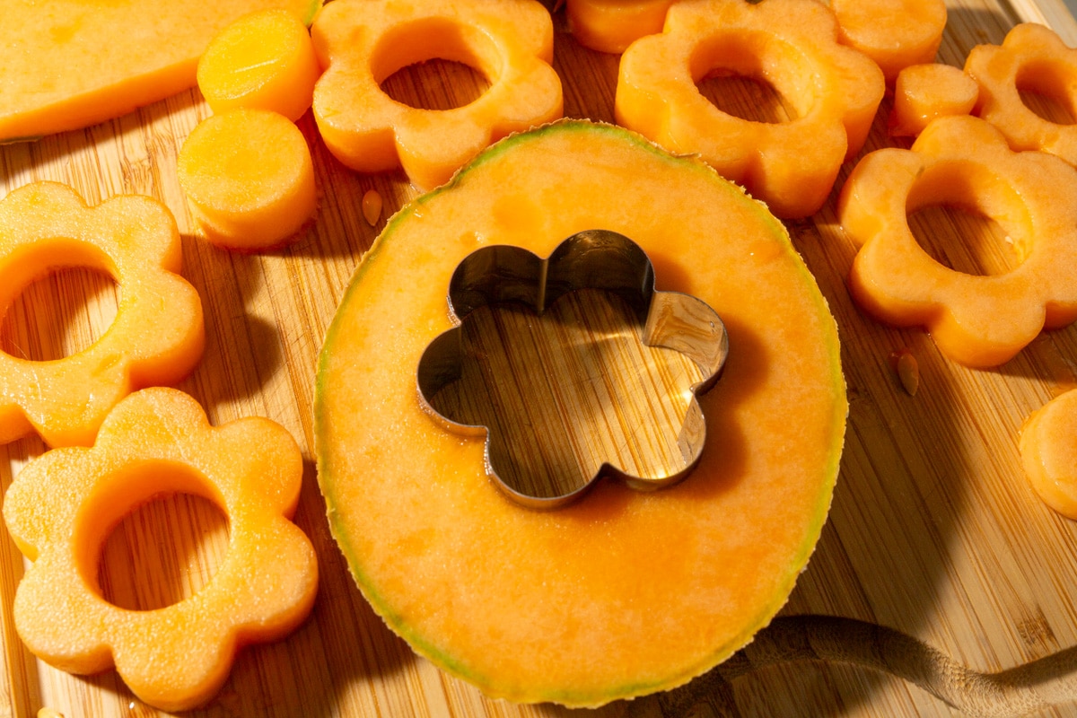 Metal flower cookie cutter pressed into a cantaloupe slice on a cutting board.