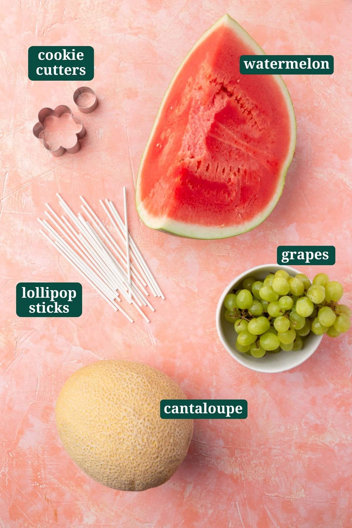 Flat lay of ingredients for flower fruit kabobs, including watermelon, cantaloupe, green grapes, cookie cutters, and lollipop sticks.