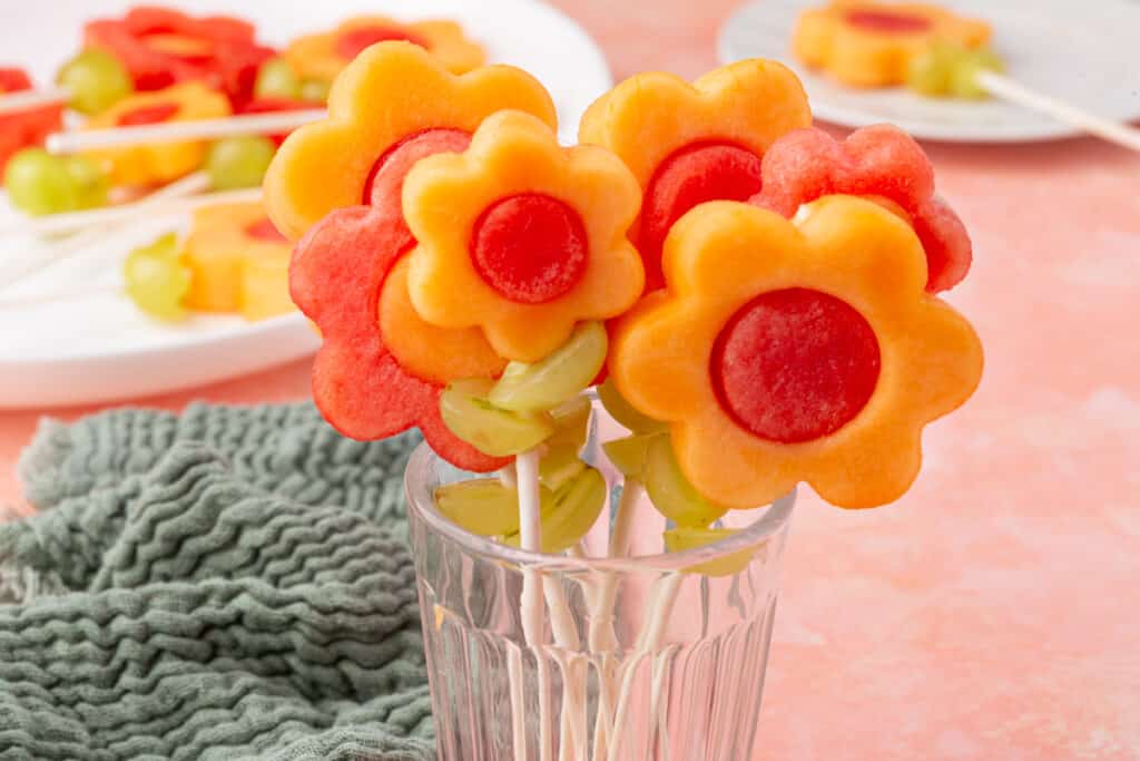 A horizontal view of fruit kabobs in a glass, with cantaloupe and watermelon flower shapes and grape accents, on a pink background.