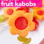 A hand holding a fruit kabob with a cantaloupe flower, watermelon center, and green grapes threaded on a stick.