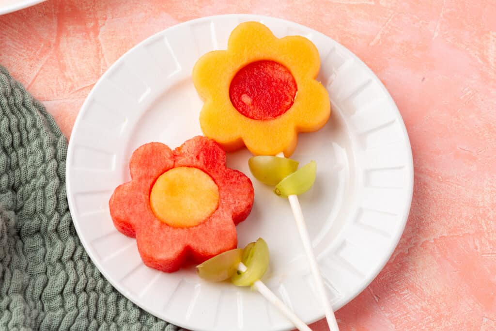 Two flower fruit kabobs on a white plate—one made with cantaloupe and watermelon, the other with watermelon and cantaloupe—each with grape leaves skewered on white sticks.