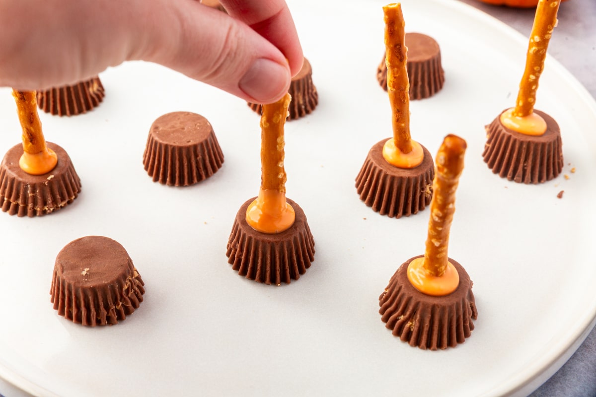 Hand pressing pretzel sticks into peanut butter cups with orange candy coating to make witch brooms.