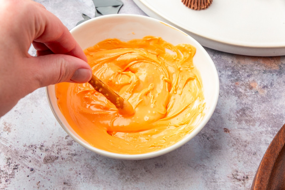 Hand dipping a pretzel stick into melted orange candy coating.