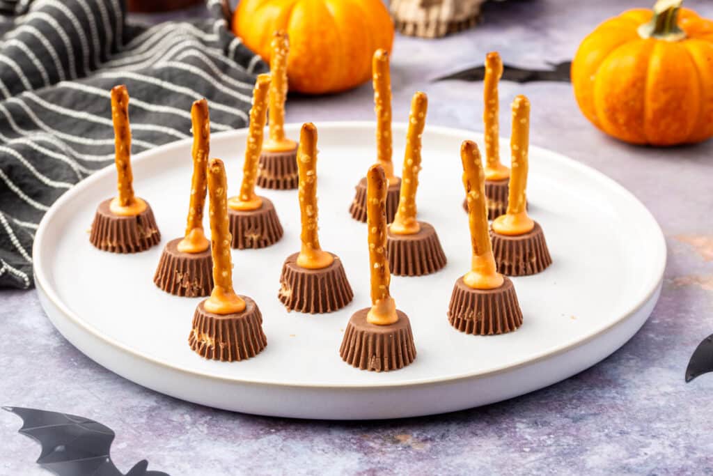 Finished peanut butter cup pretzel witch brooms standing upright on a white tray with Halloween decorations.