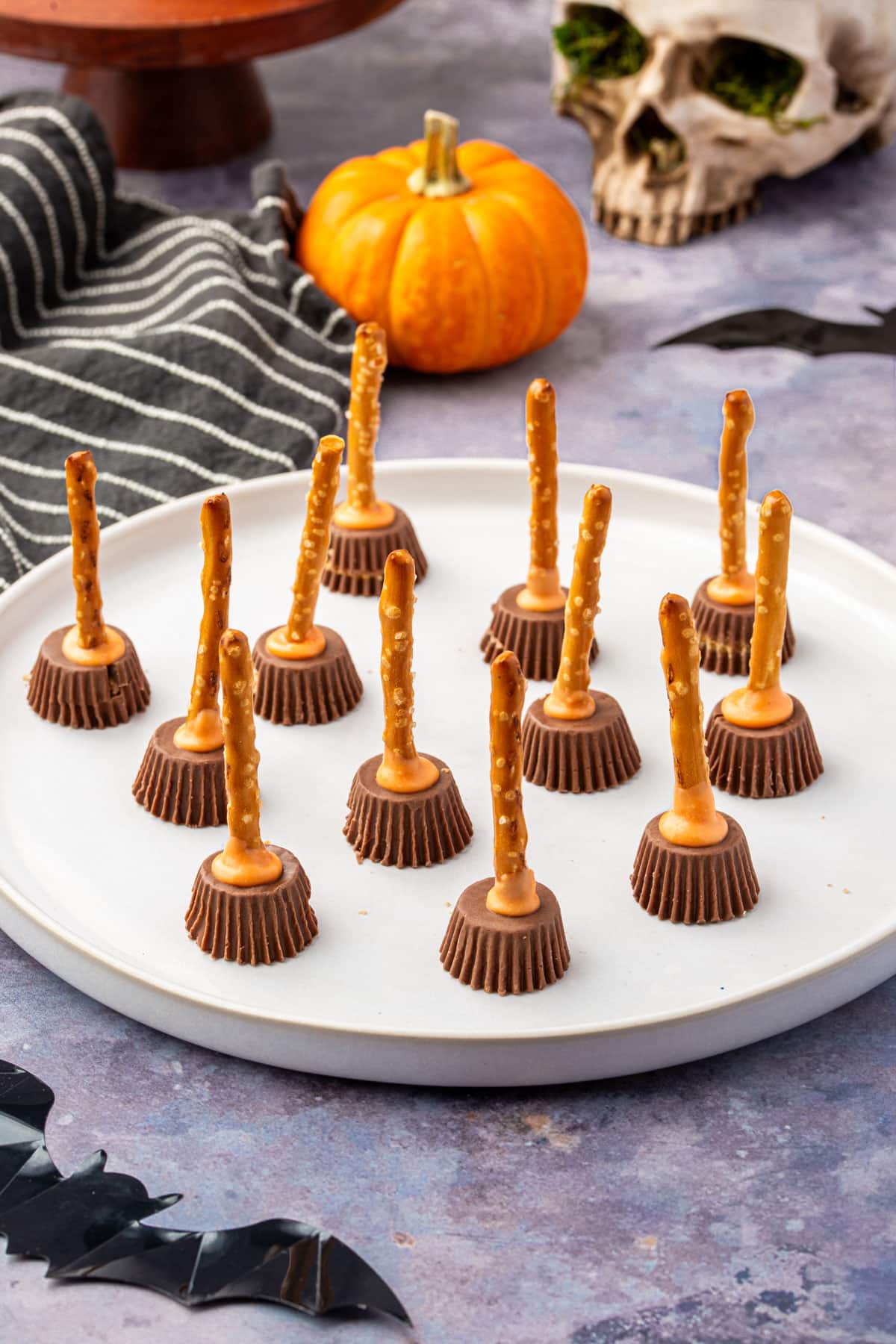 Vertical shot of peanut butter cup pretzel witch brooms on a plate surrounded by Halloween props.