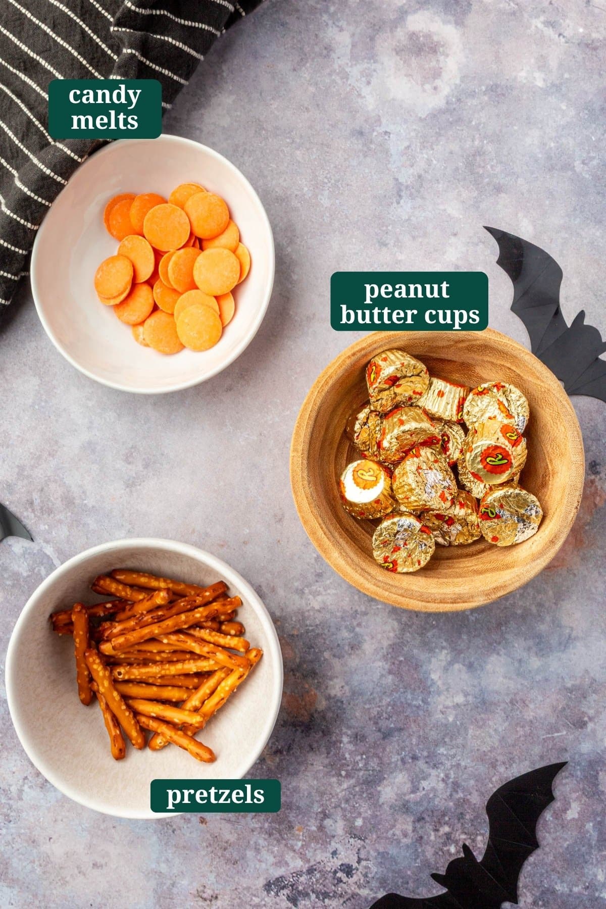 Overhead view of bowls filled with orange candy melts, pretzel sticks, and wrapped peanut butter cups for Halloween broom treats.