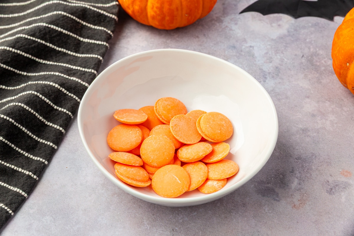 Orange candy melts in a white bowl before melting for the peanut butter cup pretzel witch brooms.
