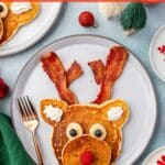 Christmas reindeer pancakes made with bacon antlers, banana eyes, and a raspberry nose on a festive holiday breakfast table.
