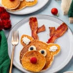 Reindeer pancake decorated with bacon antlers, banana and blueberry eyes, raspberry nose, and whipped cream ears on a festive table.