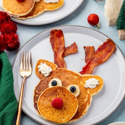 Reindeer pancake decorated with bacon antlers, banana and blueberry eyes, raspberry nose, and whipped cream ears on a festive table.