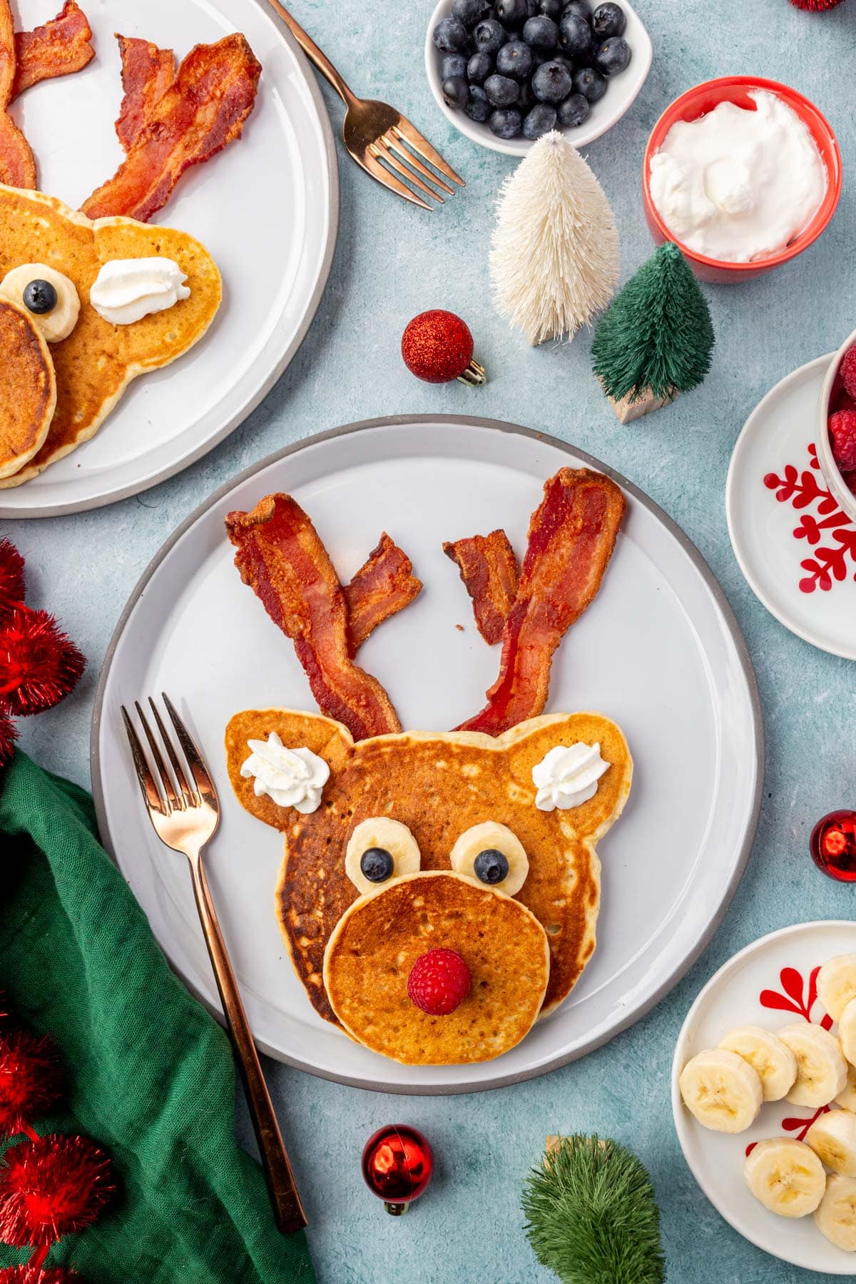 Fun holiday breakfast featuring a reindeer pancake made from two golden pancakes with bacon antlers and fruit decorations.