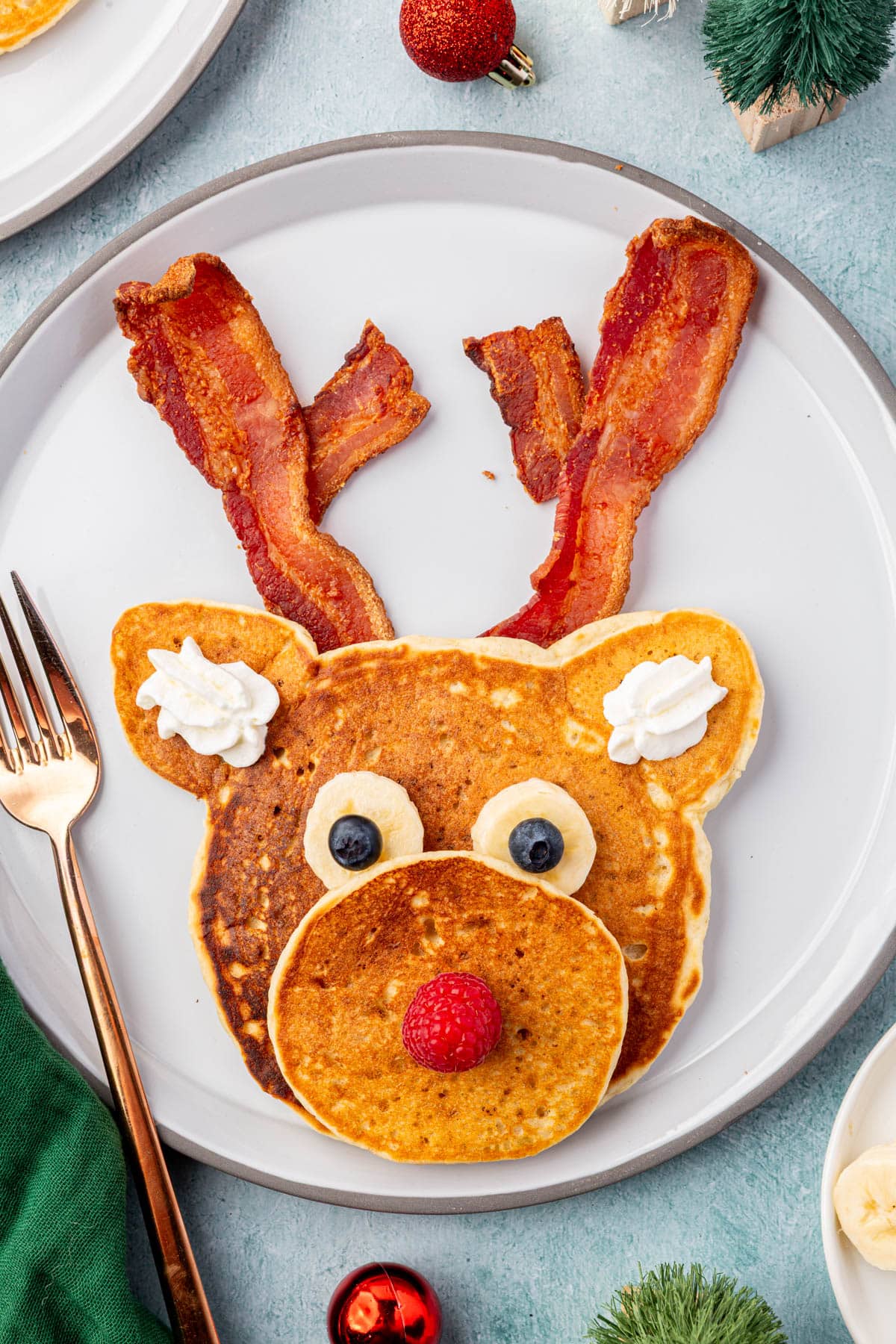 Finished reindeer pancake on a plate with bacon antlers, banana and blueberry eyes, raspberry nose, and whipped cream ears.