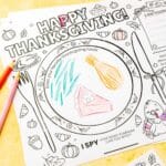Close-up of Thanksgiving placemat with "Draw your favorite Thanksgiving foods" plate section partially colored in with green beans, corn, and pink pie slice, colored pencils visible on side.