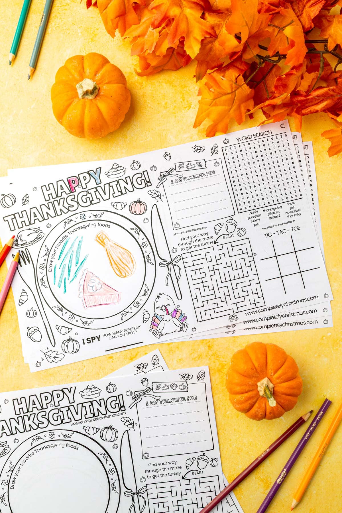 Multiple Thanksgiving activity placemats displayed with colorful pencils on yellow background with orange pumpkins and fall leaves.