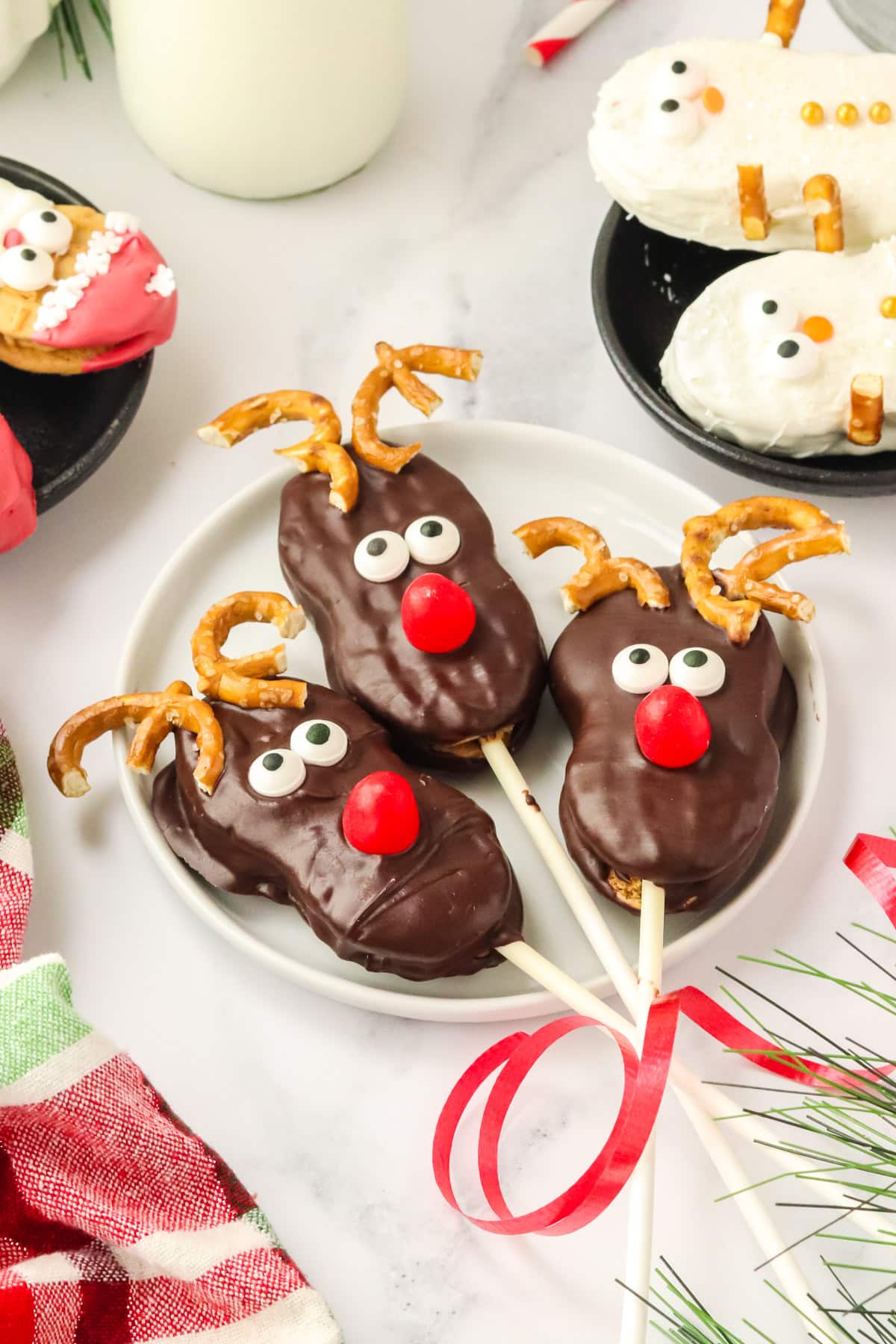 Three Nutter Butter Reindeer cookie pops on a plate with Santa and snowman cookies in the background and festive greenery.