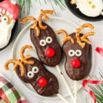 Three finished Nutter Butter Reindeer cookie pops on a white plate with pretzel antlers and red candy noses, surrounded by Santa and snowman cookie pops.