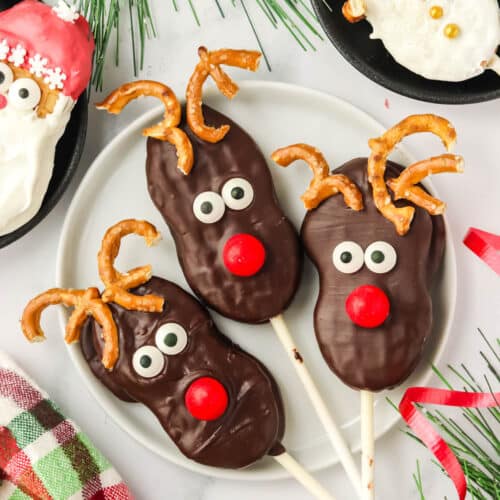Three finished Nutter Butter Reindeer cookie pops on a white plate with pretzel antlers and red candy noses, surrounded by Santa and snowman cookie pops.