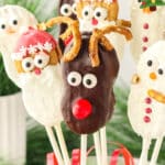 A Nutter Butter Reindeer cookie pop with pretzel antlers, candy eyes, and a red candy nose displayed with Santa and snowman cookie pops in the background.