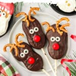 Three Nutter Butter Reindeer cookie pops on a white plate with pretzel antlers and red candy noses.