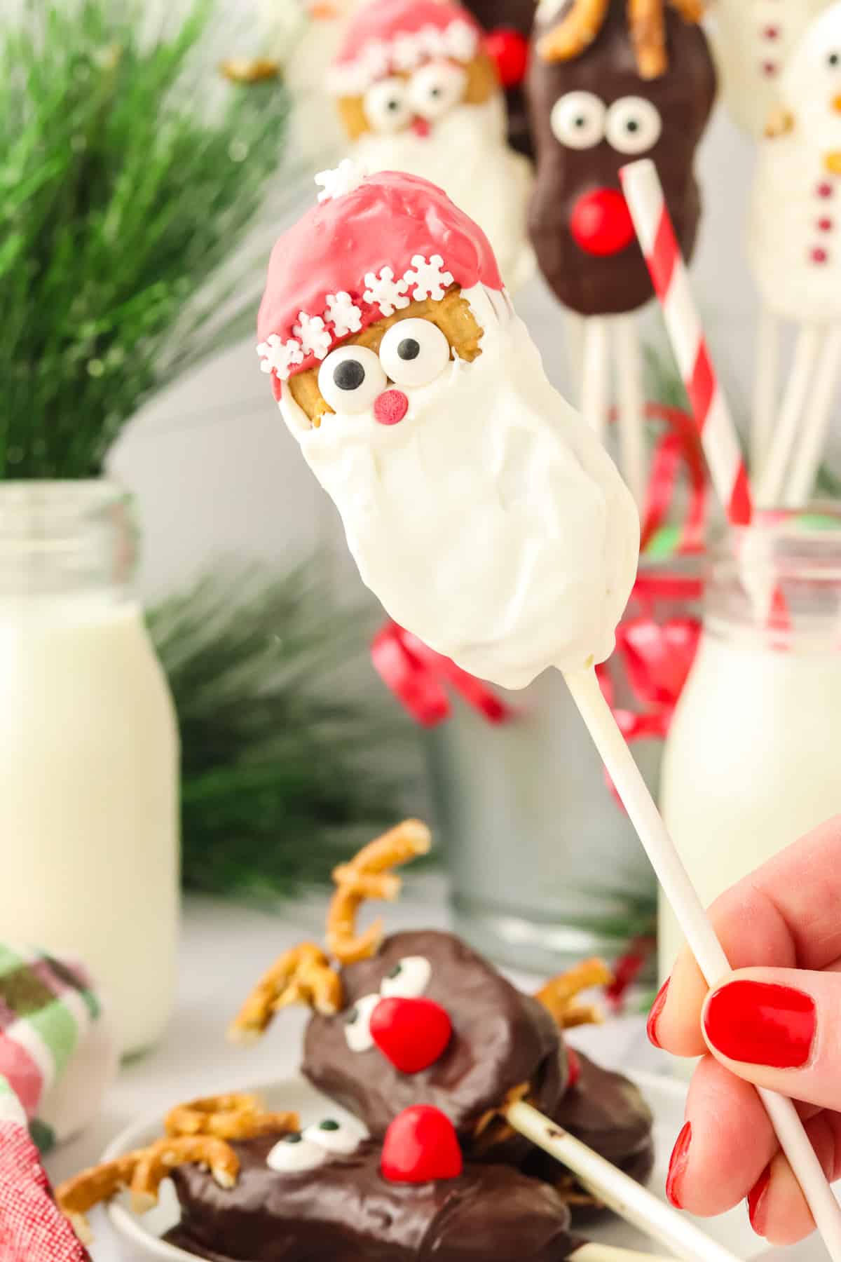 A hand holding a finished Nutter Butter Santa cookie pop with reindeer and snowman cookie pops in the background.