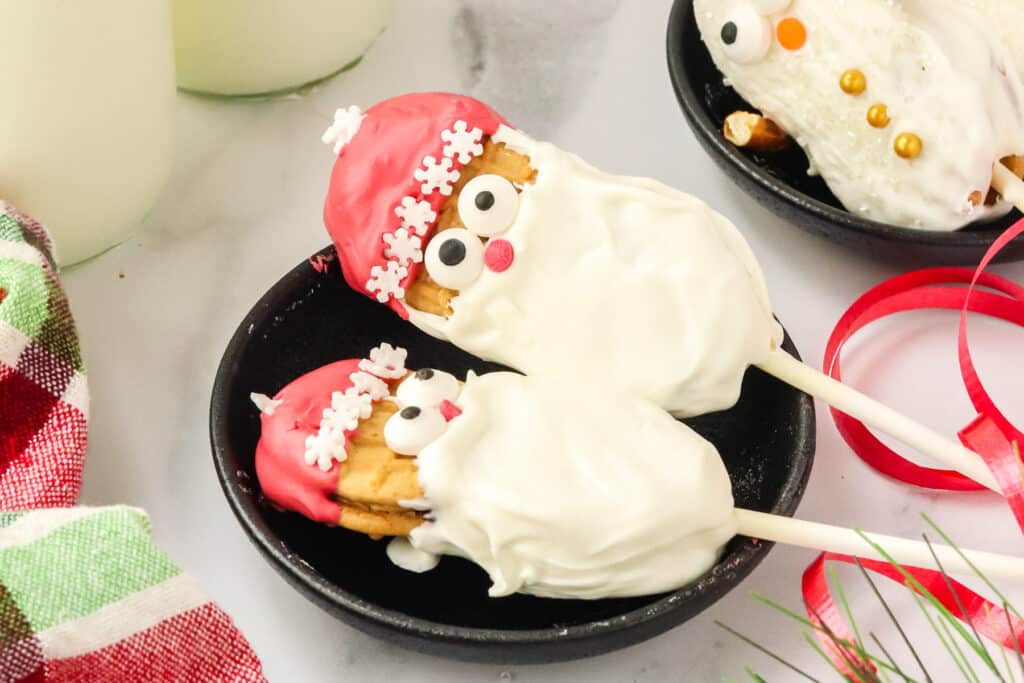 Two finished Nutter Butter Santa cookie pops with white chocolate beards, red candy hats, and snowflake sprinkle trim on a black plate.