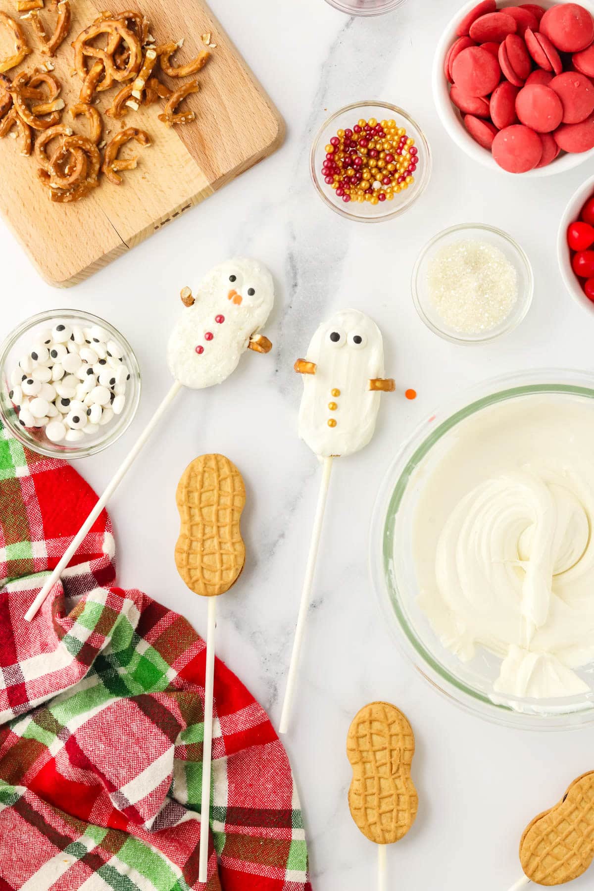 Nutter Butter Snowman cookies in progress showing one with sanding sugar and buttons and one with gold buttons being decorated.