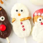 Close up of a Nutter Butter Snowman cookie pop with sanding sugar, pretzel arms, candy eyes, orange nose, and red candy buttons next to reindeer and Santa cookies.