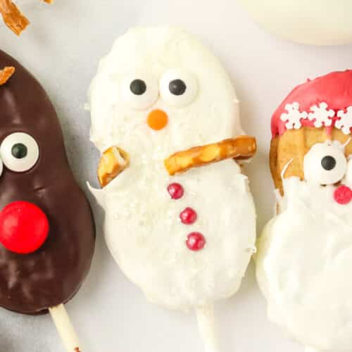 Close up of a Nutter Butter Snowman cookie pop with sanding sugar, pretzel arms, candy eyes, orange nose, and red candy buttons next to reindeer and Santa cookies.