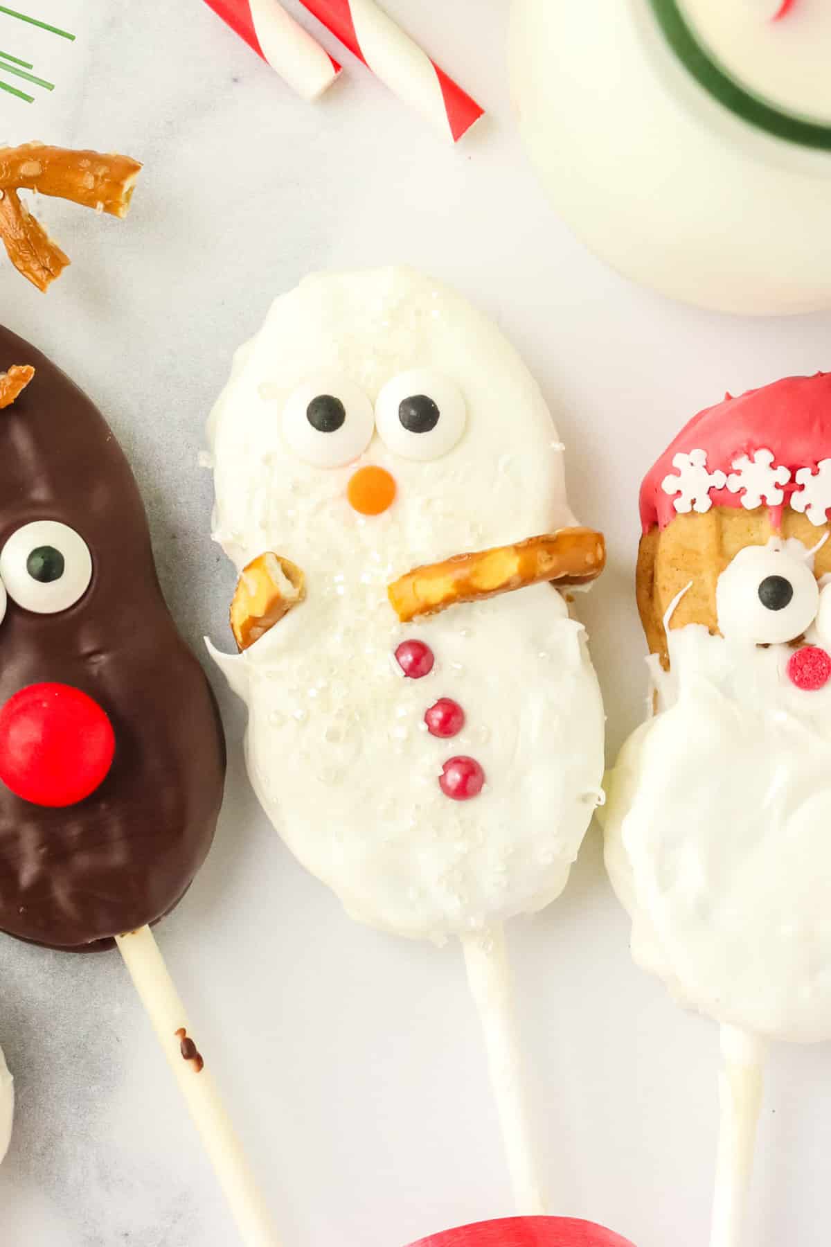 Close up of a Nutter Butter Snowman cookie pop with sanding sugar, pretzel arms, candy eyes, orange nose, and red candy buttons next to reindeer and Santa cookies.