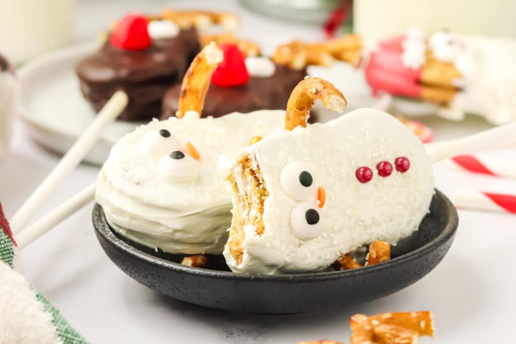 Two Nutter Butter Snowman cookie pops on a black plate with sanding sugar coating, pretzel arms, and candy decorations.