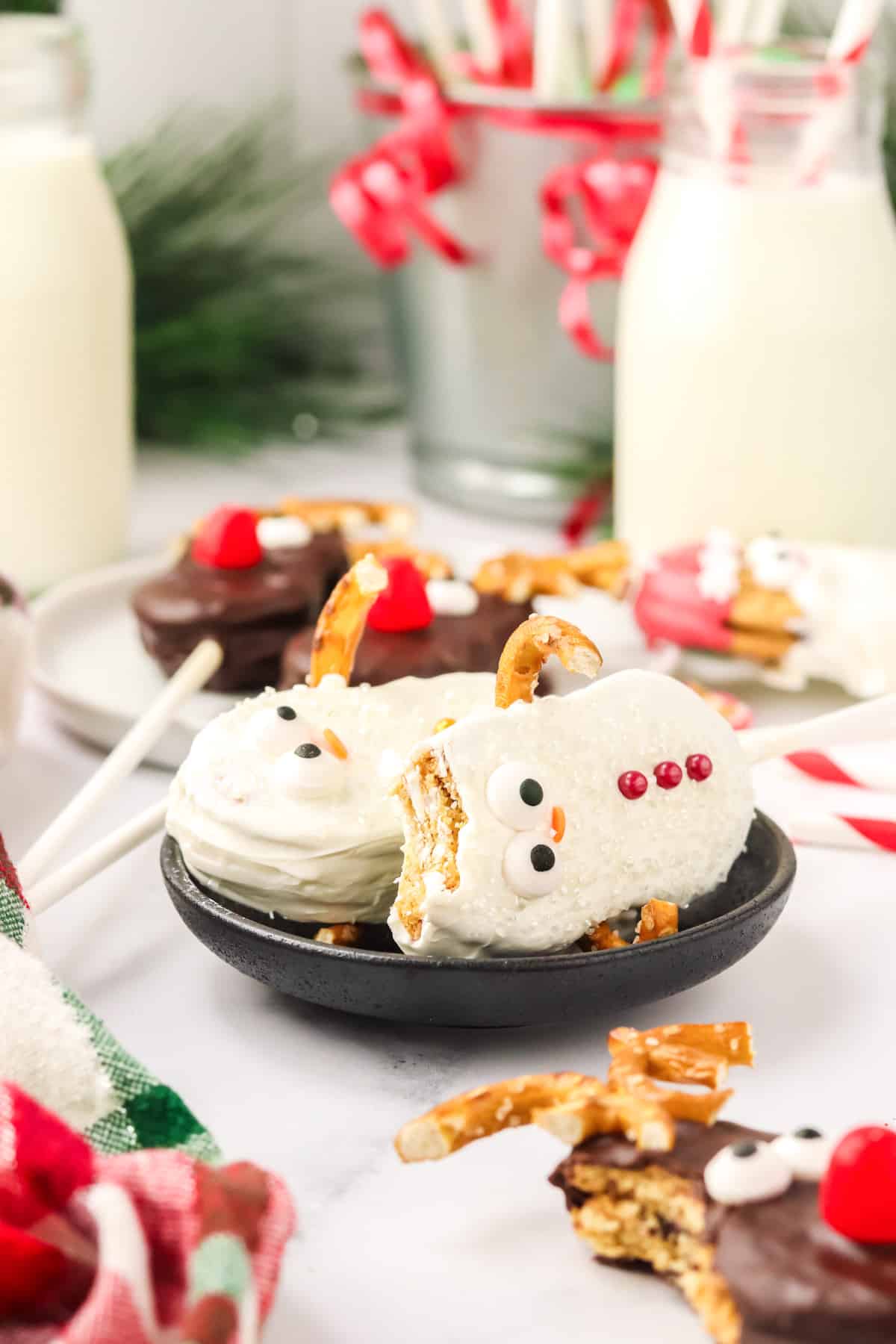 Two Nutter Butter Snowman cookie pops on a black plate with one showing a bite taken out and reindeer cookies in the background.