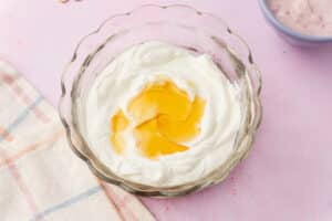 Greek yogurt with honey added to a glass mixing bowl on a pink background before stirring.