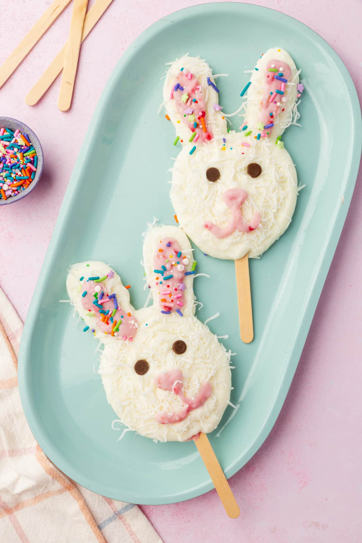 Two Easter bunny frozen yogurt pops on a blue platter decorated with coconut, sprinkles, and chocolate chip eyes.