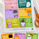 A printed Halloween lunchbox notes sheet with three cut cards fanned out in front alongside orange scissors on a light surface.