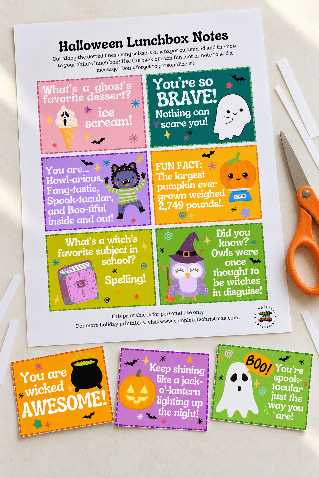 A printed Halloween lunchbox notes sheet with three cut cards fanned out in front alongside orange scissors on a light surface.